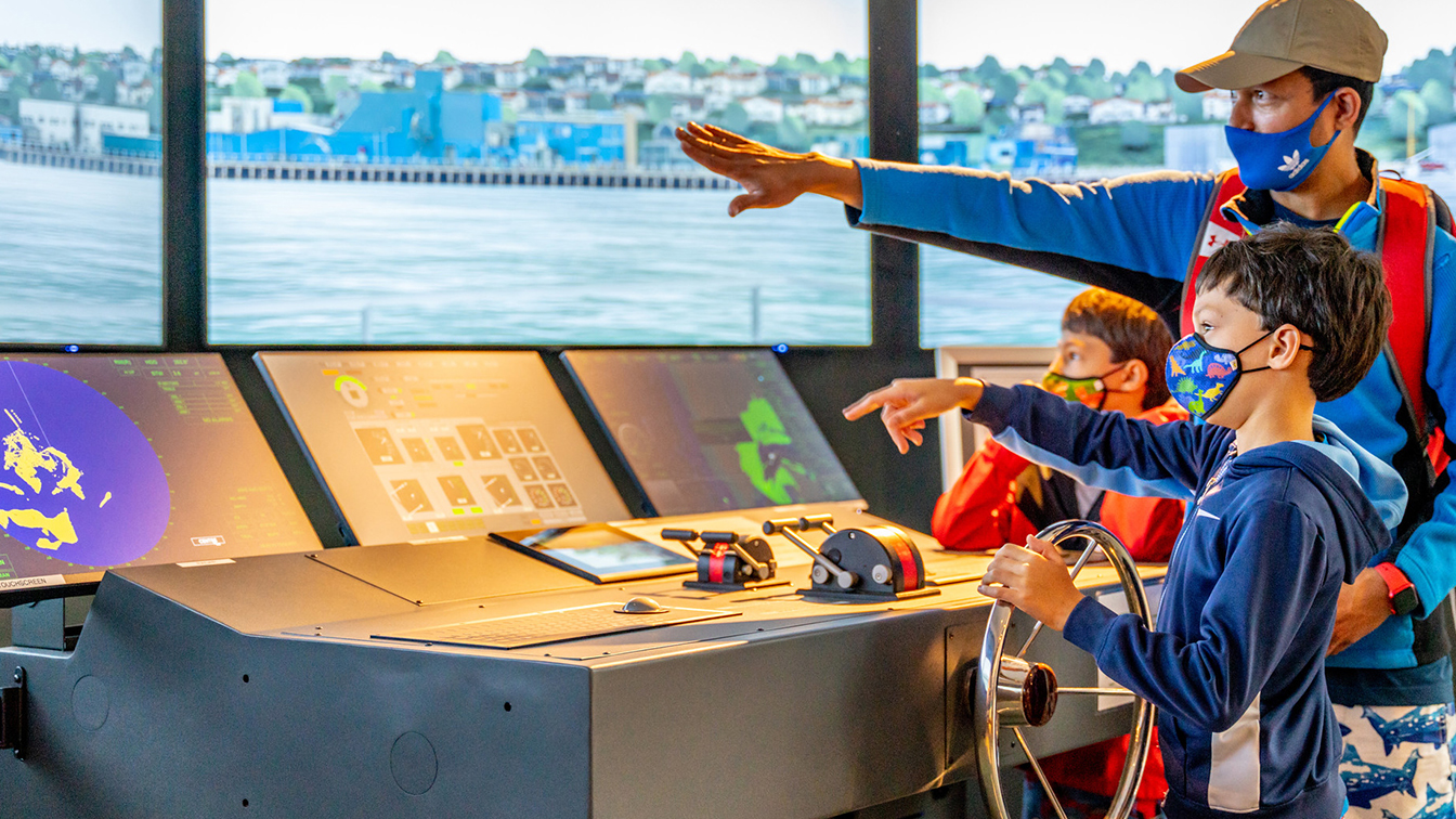 Using a simulator, a child steers a research ship through Yaquina Bay in an exhibit at the Hatfield Visitor Center. His father stands near him and points to the screen in front of them.