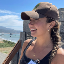 A woman in a brown Oregon State DNR baseball hat, stand on the Oregon coast and smiles into the distance.
