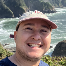 A man in a baseball style hat smiles into the camera. In the background is the Pacific Northwest coastal shoreline and cliffs.
