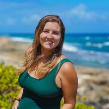 Jess Schulte stands near the beach on a sunny day. The ocean is in the background.