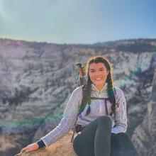 Rebecca Smoak sits on a moutain top with a view of a canyon in the background.