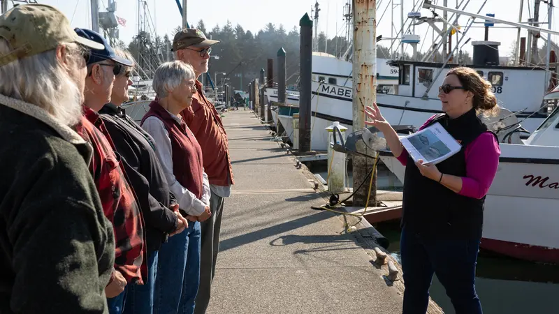 A person gestures to a group of people on a dock in front of boats.
