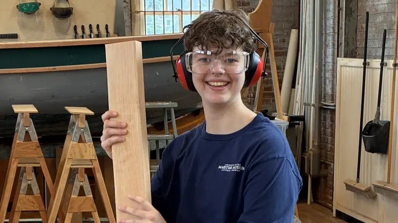 A person wearing safety goggles and holding a plank of wood smiles at the camera.