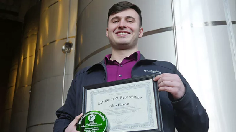 Alan Haynes, former OSU student and intern at Widmer Brothers Brewing, stands in front of brewery tanks holding award for pollution prevention