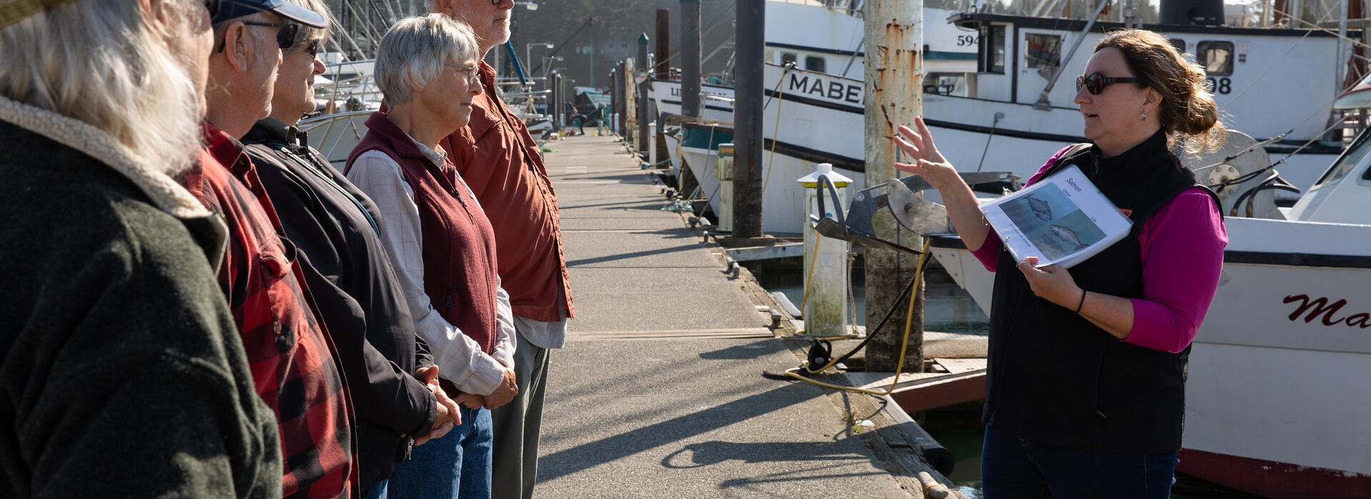 A person gestures to a group of people on a dock in front of boats.