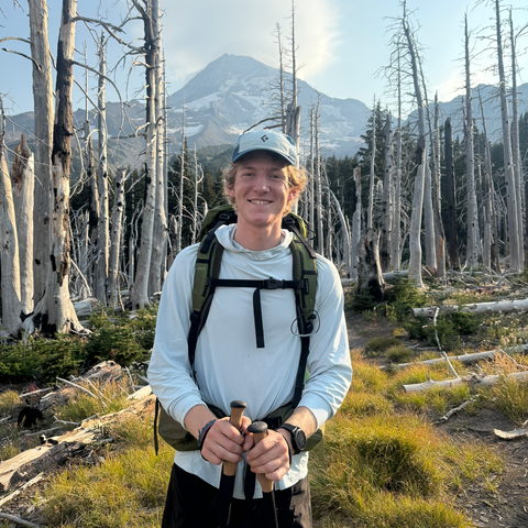 A person smiles at the camera in front of a mountain.