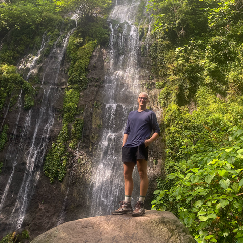 A person smiles at the camera in front of a waterfall.