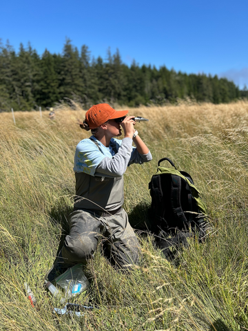 A person looks through binoculars while kneeling in the grass.
