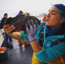 A woman in yellow bib wadders, holds a rockfish up to her face as if to give it a kiss. 