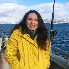 A woman with long black hair and wearing a yellow rain coat stands on a boat and smiles at the camera. A fishing rod is close by and the background shows snow covered mountains at the shoreline.