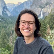 A young woman with shoulder length brown hair and classes smiles into the camera. The background of this photo show the mountain valley with a waterfall. 